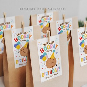 Op de afbeelding: Verschillende bruine papieren cadeauzakjes met houten knijpers die labels vasthouden. De labels lezen "Chip Chip Hooray, it's my Birthday!" met een cartoon koekjespersonage, ballonnen en een feesthoed.