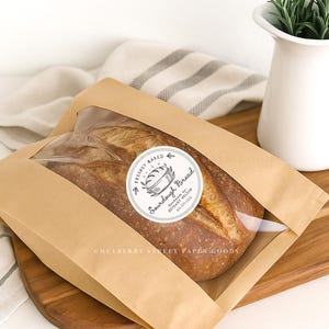 Puede incluir: Una hogaza de pan de masa madre reci&eacute;n horneado en una bolsa de papel marr&oacute;n con una ventana transparente. La bolsa tiene una pegatina redonda que dice "Freshly Baked Sourdough Bread". El pan est&aacute; sobre una tabla de cortar de madera.