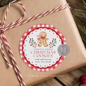 Puede incluir: Un regalo envuelto con una cinta roja y blanca. Una pegatina redonda con la inscripción "FRESHLY BAKED CHRISTMAS COOKIES" con una ilustración de hombre de jengibre. La pegatina tiene un borde a cuadros rojo y blanco y un borde festoneado.