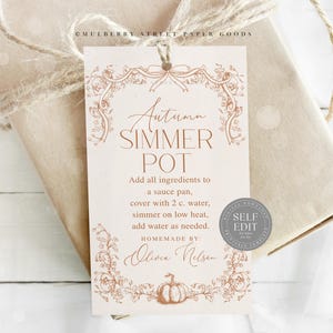 May include: A beige gift tag with the text "Autumn Simmer Pot" in elegant script, adorned with floral and pumpkin illustrations. The tag includes instructions and is tied to a wrapped gift with twine.