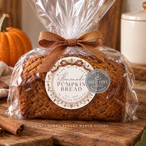 Puede incluir: Una hogaza de pan de calabaza empaquetada en una bolsa de plástico transparente con un lazo de cinta marrón. Una etiqueta circular en el frente presenta el texto "Made with love", "Homemade PUMPKIN BREAD", "Nov. 20XX" y "by Allison Thompson" con un borde de hojas.