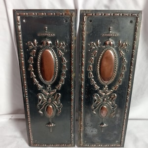 Peut inclure: Une paire de plaques de porte rectangulaires en métal avec une finition sombre et des accents cuivrés. Chaque plaque présente un centre ovale en relief, des volutes décoratives, un motif de nœud et un petit détail ovale, bordés d'une frise perlée.