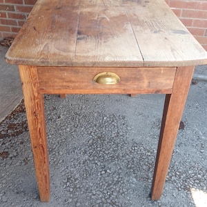 May include: A rectangular wooden table with a natural brown finish and visible wood grain. The table features a single drawer centred on the front apron, equipped with a semi-circular brass cup pull handle. The four legs are straight and tapered.