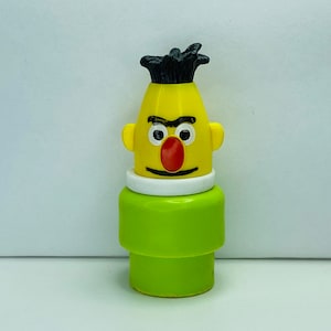 Pode incluir: Uma figura de brinquedo de plástico amarelo com uma peça de cabelo preto, um nariz vermelho e uma base verde. A figura tem uma expressão de raiva.