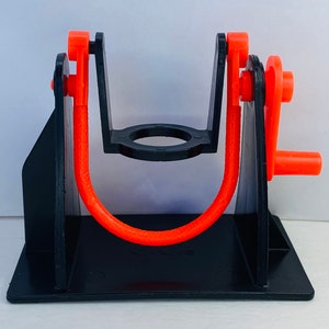 May include: A black plastic device with an orange rubber band and a handle. The device is used to stretch and exercise the fingers.
