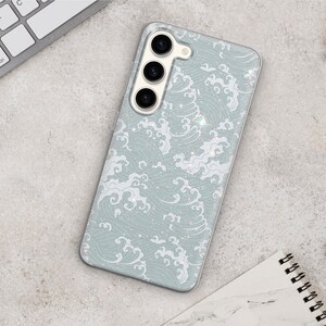 Puede incluir: Una funda transparente para tel&eacute;fono con un patr&oacute;n de olas azules y blancas y detalles de purpurina. La funda tiene un recorte para una lente de c&aacute;mara triple.