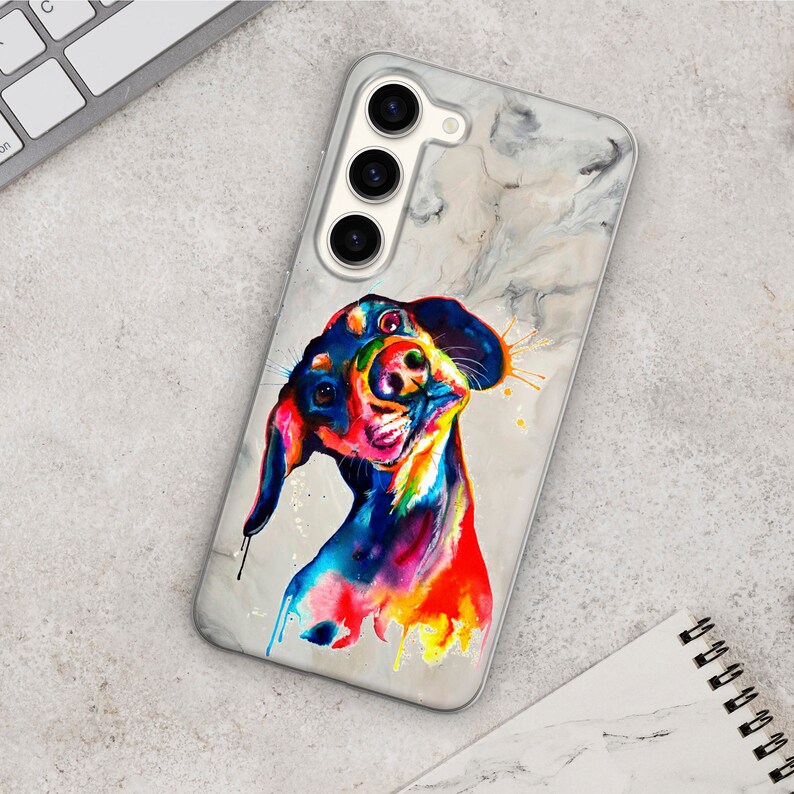May include: A white phone case with a marble pattern and a colorful abstract painting of a dachshund dog. The dog is painted in shades of blue, green, yellow, orange, and red.