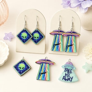 May include: A collection of alien-themed jewelry featuring two pairs of dangle earrings and three loose charms. The designs include green alien faces on blue star-patterned diamonds, colorful UFOs with tractor beams, and a UFO with the text Take Me Away.