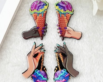 May include: Two pairs of dangle earrings. One pair features a hand holding a purple, green, and yellow ice cream cone. The other pair features a hand holding a blue, green, and purple galaxy orb.
