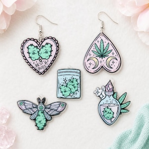 May include: A set of five acrylic charms and earrings featuring pastel-colored designs. Includes a heart-shaped earring with the text BEST BUDS, a spade-shaped earring with a leaf, a rectangular baggie, a moth, and a potion bottle, all with green buds.