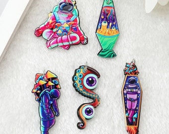 May include: A collection of colorful acrylic charms featuring space and psychedelic themes. Designs include an astronaut, a lava lamp, an eye with tentacles, and an astronaut in a coffin. Each charm has a silver-colored loop for hanging.