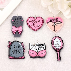 May include: Six pink and black enamel charms with feminist slogans. The charms are shaped like a coffin, a heart, a bra, a mirror, and two tombstones. The text on the charms includes "RIP beauty standards", "Thick thighs save lives", "Badass babe", "Burn your bras", "RIP gender roles", and "I see a powerful woman".