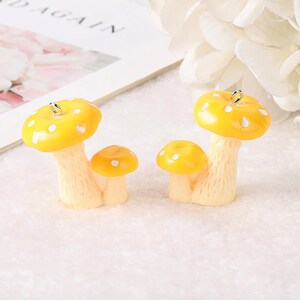 May include: Two yellow mushroom charms with white spots on the caps. Each charm features a larger mushroom and a smaller mushroom attached to a textured cream-colored stem. A small silver metal loop is attached to the top of each larger cap.