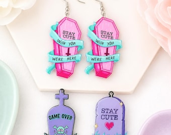 May include: A pair of pink coffin-shaped earrings with teal ribbons and the text STAY CUTE WISH YOU WERE HERE, plus two tombstone-shaped charms. One purple tombstone features a skull and GAME OVER, while the other has a heart and STAY CUTE.