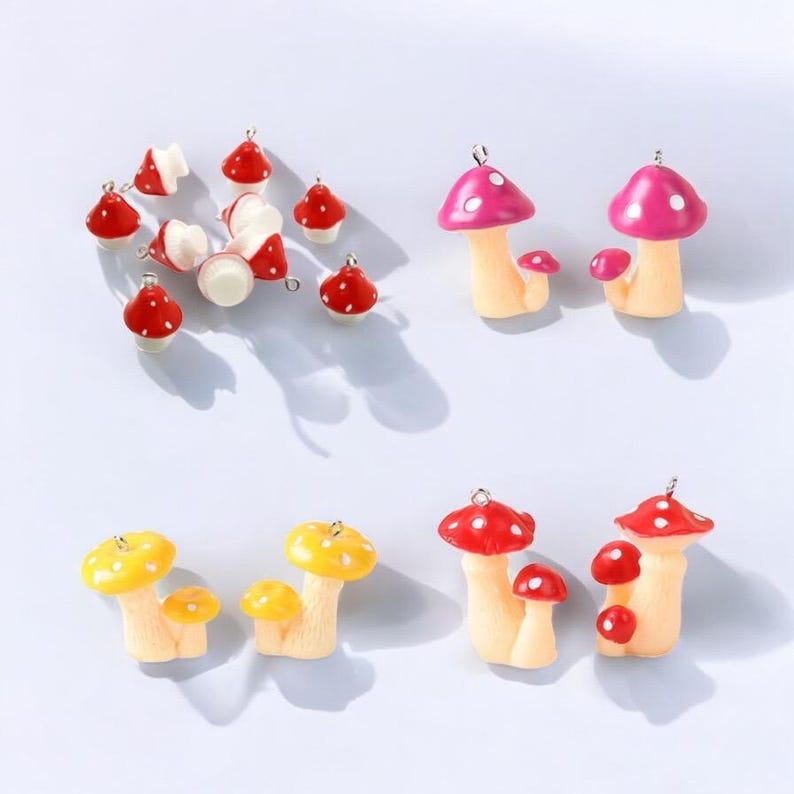May include: A collection of small mushroom charms in various colors including red, pink, and yellow. Each charm features a cap with white spots and a light-colored stem, equipped with a small metal loop at the top for attachment to jewelry.