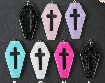 May include: Eight coffin-shaped charms with a cross cutout. The charms are made of resin and have glitter embedded in them. The colors of the charms are black, pink, purple, light blue, white, hot pink, and black.