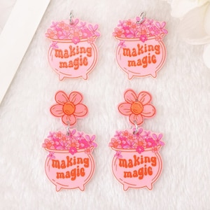 May include: Pink and orange acrylic earrings with a flower charm and a cauldron charm. The cauldron charm has the text "making magic" on it.