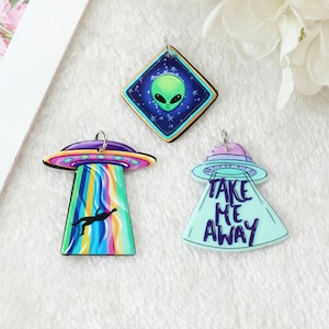 May include: Three colorful alien-themed pendants. One pendant features a green alien head on a blue starry background. Another shows a UFO with a rainbow beam and a silhouette. The third pendant says "TAKE ME AWAY".