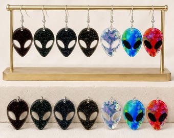 May include: A collection of seven alien-shaped earrings and seven matching pendants. The designs include black glitter, iridescent, blue and green, and red and orange speckled patterns. Each piece features a cutout eye design and silver hardware.