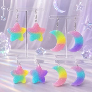 May include: A collection of star and crescent moon shaped earrings featuring a glittery, multi-colored gradient finish. The set includes two pairs of dangling star earrings and two pairs of crescent moon earrings, all suspended from silver-toned hooks.