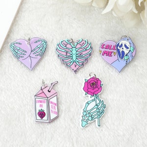 May include: Five pink and blue heart-shaped charms with different designs. One charm features a skeleton hand holding a heart, another has a skeleton ribcage, another says "Call Me" with a skeleton wearing a ghost mask, another has a strawberry milk carton with the text "Don't Drink Real Blood", and the last charm has a skeleton hand holding a pink rose.