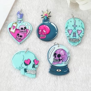 May include: Five colorful charm pendants featuring skull designs. The charms are made of acrylic and have a glossy finish. The designs include a skull with hearts for eyes, a skull in a snow globe, a skull with a cross, a skull with a bomb, and a skull in a potion bottle.