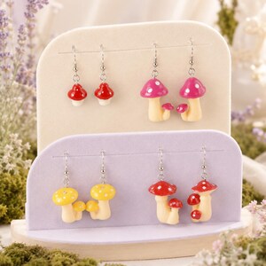 May include: Four pairs of dangle earrings featuring mushroom charms in various colors. The top row includes small red mushrooms and larger pink mushrooms with white spots. The bottom row features yellow mushrooms and red mushrooms with multiple caps.