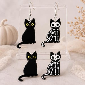 May include: Two pairs of cat-shaped earrings featuring a black cat with yellow eyes on one side and a white skeleton print on the other. The earrings are suspended from silver-toned metal hooks, showcasing a distinct contrast between the solid black feline silhouette and the detailed skeletal structure visible on the matching pair.
