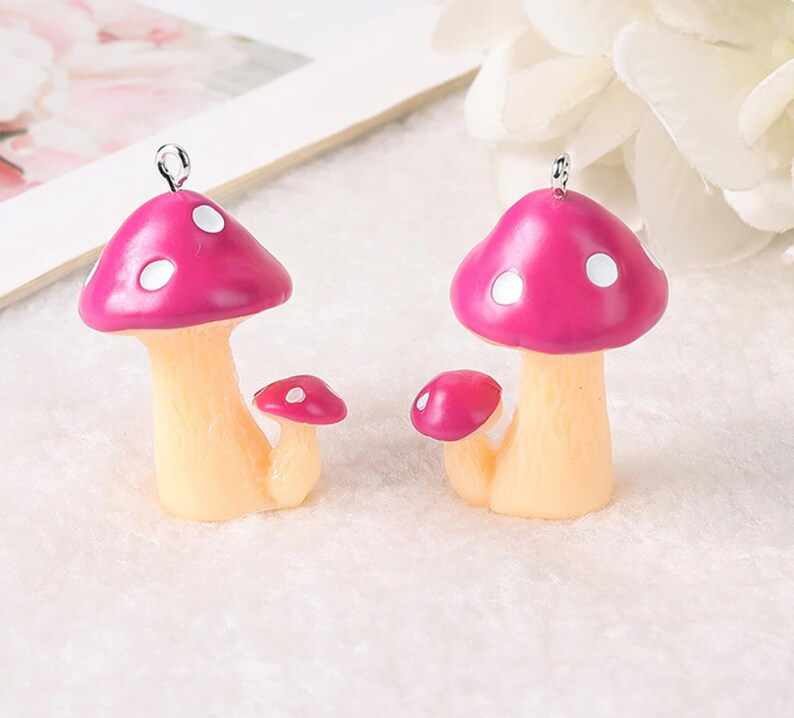 May include: Two mushroom charms featuring bright pink caps with white spots and pale beige stems. Each charm includes a smaller secondary mushroom attached to the base and a small silver metal loop at the top for hanging or attaching to jewelry.