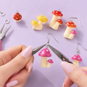 May include: A pair of metal pliers holding a pink mushroom earring with white spots. Other mushroom charms and earrings in red, yellow, and pink are scattered nearby, along with additional earring hooks and jump rings on a light purple surface.