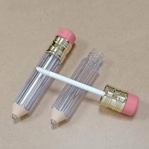 May include: Two clear plastic pencil-shaped lip gloss tubes with gold accents and pink eraser tops. The tubes are empty and have a ribbed texture. One tube has a white applicator wand.