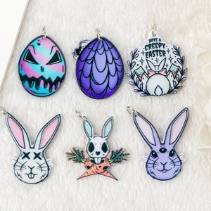 May include: Six colorful Easter-themed charms featuring a purple and blue egg with a dragon design, a purple and blue egg with a skull and crossbones design, a white bunny with a skull and crossbones design, a white bunny with a pink bow and the text "Have a Creepy Easter", a white bunny with X's for eyes, and a purple bunny with three eyes.
