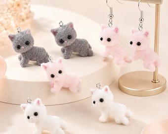 Puede incluir: Una colección de dijes con forma de gato difuso en gris, rosa y blanco. Dos dijes de gato rosa están unidos a ganchos de aretes plateados que cuelgan de un soporte de metal dorado. Cuatro dijes de gato adicionales están sobre una base circular.