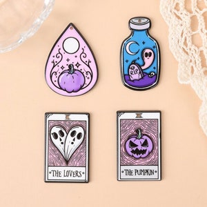 May include: Four Halloween-themed charms. Includes a planchette with a pumpkin, a bottle with a ghost, and two tarot cards. The tarot cards read "The Lovers" and "The Pumpkin". The charms are purple, black, and white.