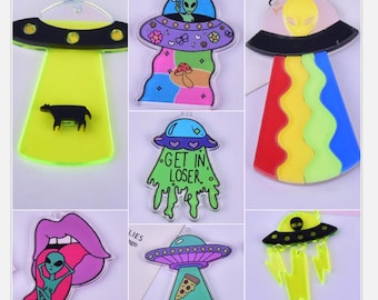 May include: Set of 8 colorful acrylic charms featuring alien and UFO designs. The charms include a cow, a pizza, a rainbow, a dripping green alien, a tongue with an alien, and a lightning bolt.