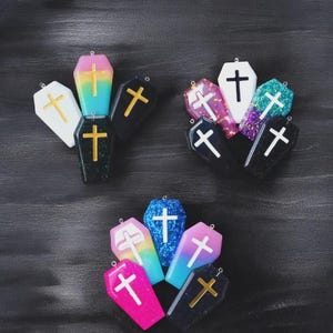 May include: A collection of colorful coffin-shaped pendants. The pendants feature glitter and crosses in various colors, including white, gold, and rainbow gradients. The pendants are arranged on a dark background.