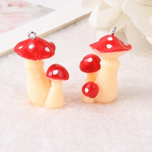 May include: Two mushroom charms featuring red caps with white spots and cream-colored stems. Each charm has a small silver metal loop at the top for attachment. The mushroom figures are sculpted with textured stems and smooth, rounded caps.