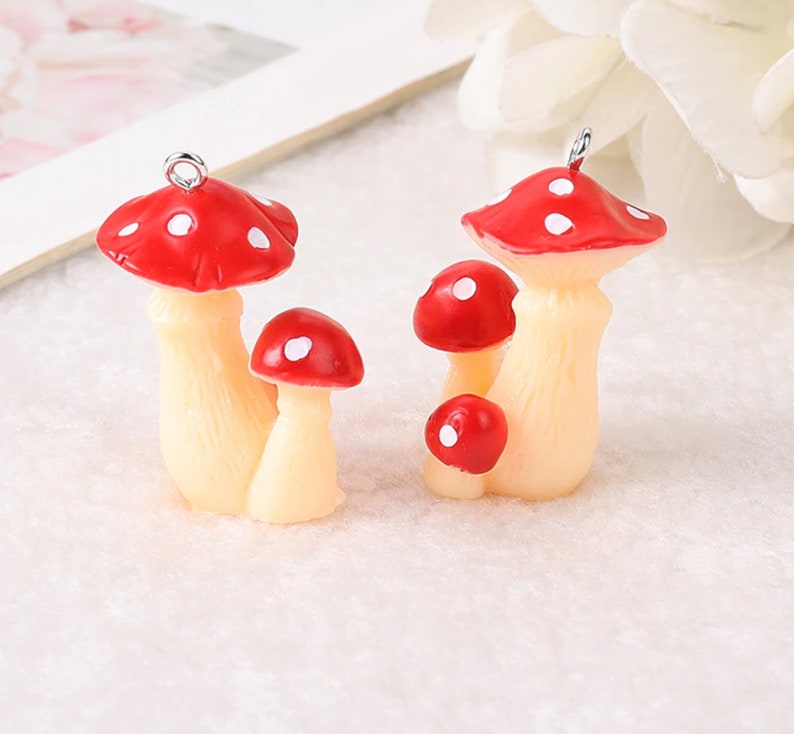May include: Two mushroom charms featuring red caps with white spots and cream-colored stems. Each charm has a small silver metal loop at the top for attachment. The mushroom figures are sculpted with textured stems and smooth, rounded caps.