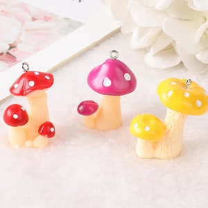 May include: Three mushroom charms with silver-tone metal loops at the top. One charm is red with white spots, one is magenta with white spots, and one is yellow with white spots. Each charm features a larger mushroom and a smaller mushroom base.