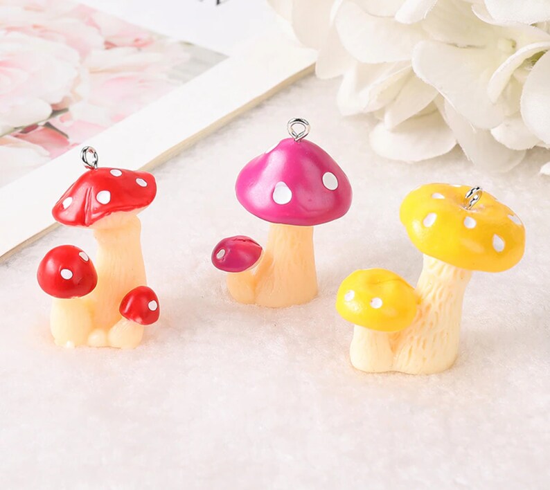 May include: Three mushroom charms with silver-tone metal loops at the top. One charm is red with white spots, one is magenta with white spots, and one is yellow with white spots. Each charm features a larger mushroom and a smaller mushroom base.