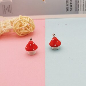 May include: Two small mushroom charms featuring bright red caps with white polka dots and white stems. Each charm includes a small silver-toned metal loop at the top for attachment, designed for use in jewelry making or as decorative craft components.