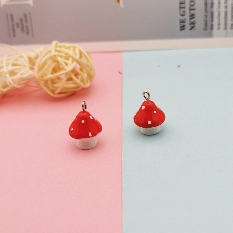 May include: Two small mushroom charms featuring bright red caps with white polka dots and white stems. Each charm includes a small silver-toned metal loop at the top for attachment, designed for use in jewelry making or as decorative craft components.