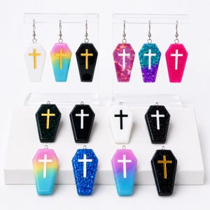 May include: A collection of coffin-shaped earrings and pendants featuring various colors and finishes. The pieces include solid white, black, pink, and blue, as well as glittery, gradient, and metallic textures, each adorned with a central cross.