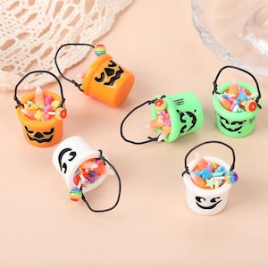 May include: Five miniature Halloween buckets filled with candy. The buckets are orange, green, and white and feature jack-o-lantern faces. Each bucket has a handle and a small lollipop attached.