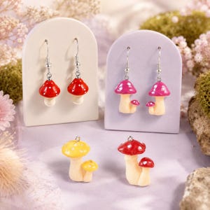 May include: A collection of mushroom-shaped jewelry pieces including two pairs of drop earrings and two individual charms. The earrings feature red and pink mushroom caps with white spots on silver hooks. The charms are yellow and red mushrooms.