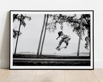 May include: Black and white photograph of a person skateboarding in front of palm trees. The person is wearing a cap and is in mid-air, performing a trick.