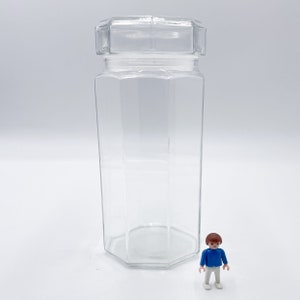 Puede incluir: Un tarro de vidrio transparente, octogonal, con tapa a juego. El tarro es alto y delgado, con un diseño geométrico. Una pequeña figura de juguete azul y blanca está al lado del tarro.