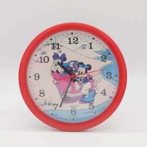 May include: Red plastic clock featuring Mickey Mouse and Minnie Mouse in a pink car against a beach backdrop. The clock face has black numbers and hands. The clock reads "Disney by Avon".