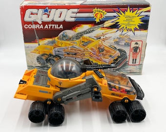 May include: An orange and black G.I. Joe Cobra Attila toy vehicle with a clear dome and multiple wheels. The box behind the toy features illustrations of the vehicle and the text "G.I. Joe Cobra Attila".