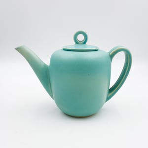 Puede incluir: Una tetera de cerámica de color verde azulado con un cuerpo redondeado y una simple asa en forma de anillo. La tetera tiene un acabado liso y esmaltado.